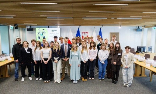 Grupa elegancko ubranych uczniów z nauczycielem stoi w sali.